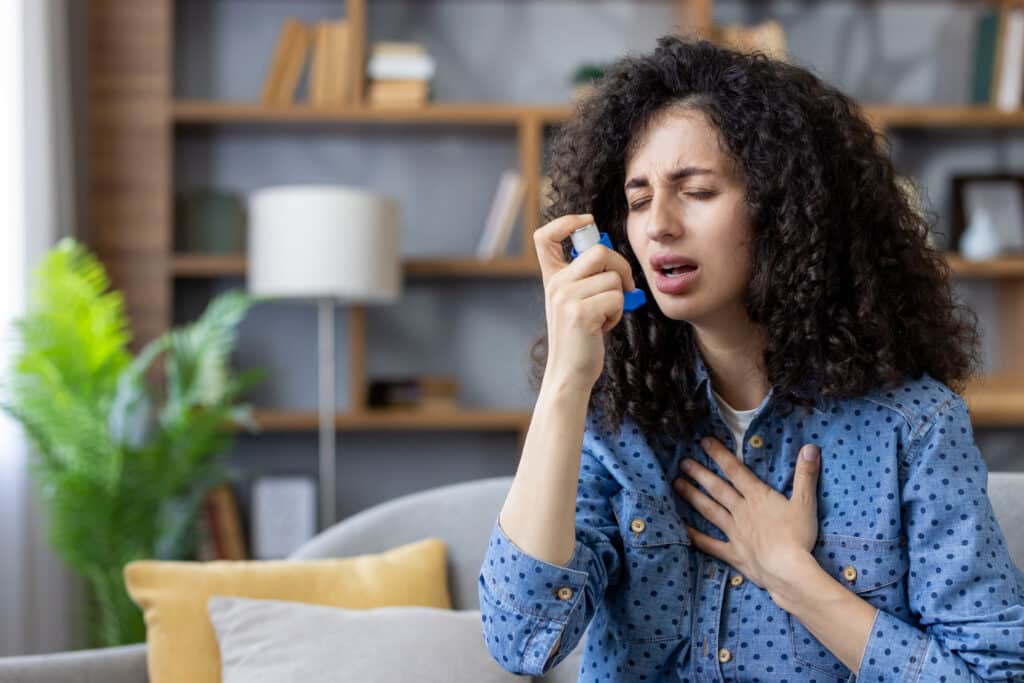 Woman using an inhaler at home with a hand on her chest